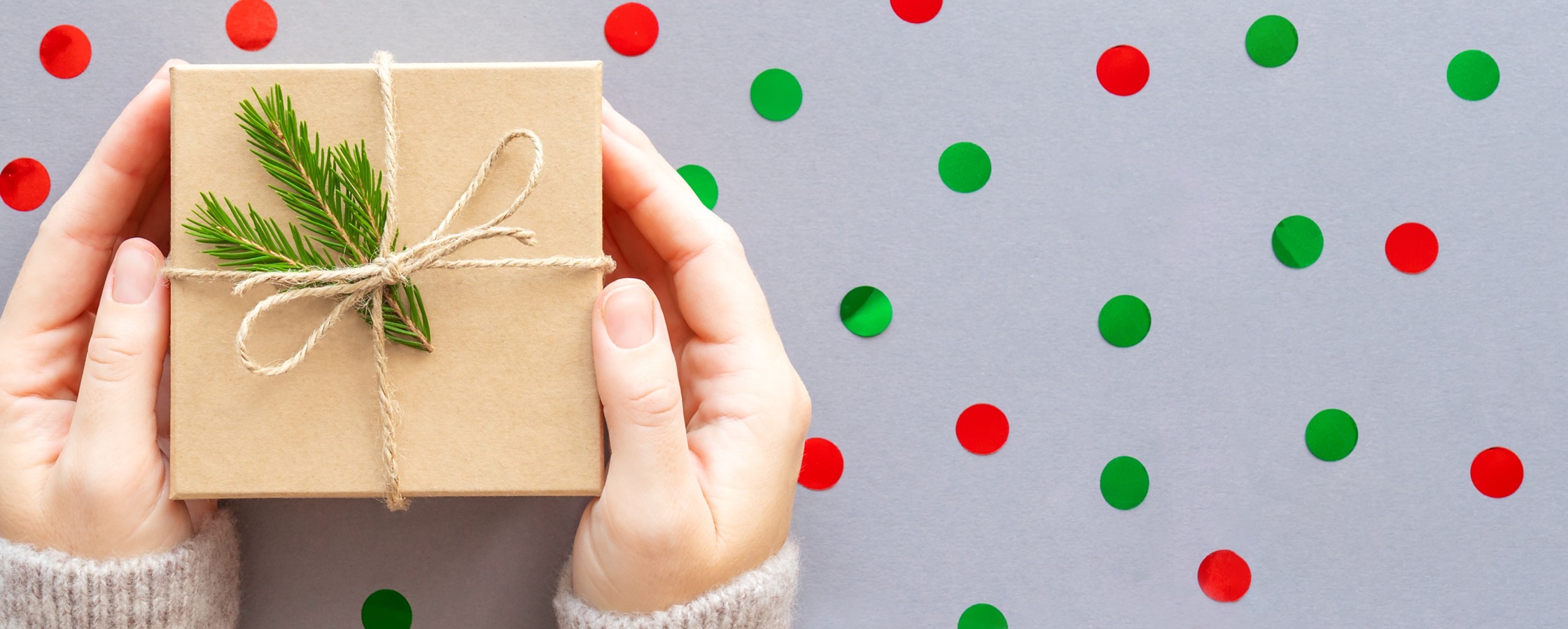 Två händer håller ett inslaget paket med snöre och grankvist mot en bakgrund med röda och gröna prickar.