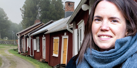 En person med blå halsduk står framför röda trähus med vita knutar på en grusväg omgiven av grönska.