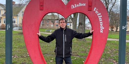 Man står i en stor röd hjärtformad ram i en park med träd och hus i bakgrunden.