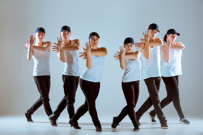 Sex personer dansar i synkroniserad formation, klädda i vita linnen, svarta byxor och kepsar.