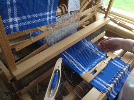 En person väver blått och vitt tyg på en vävstol nära ett fönster.