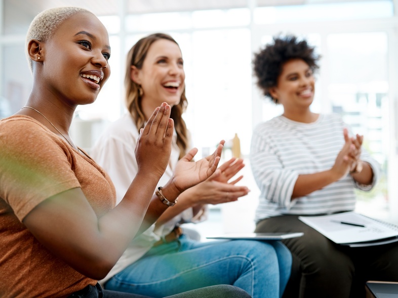 Tre kvinnor sitter tillsammans inomhus och applåderar, ser glada och engagerade ut.