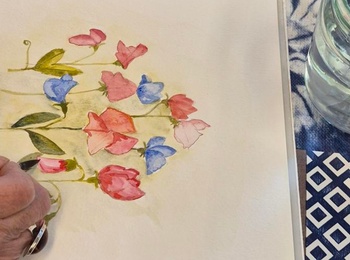 En person målar blommor med akvarellfärger vid ett bord fullt av konstmaterial.