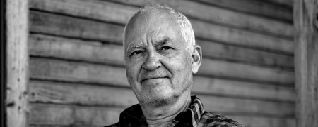 Äldre man med kort hår sitter på en veranda framför en trävägg, bär rutig skjorta. Svartvit bild.