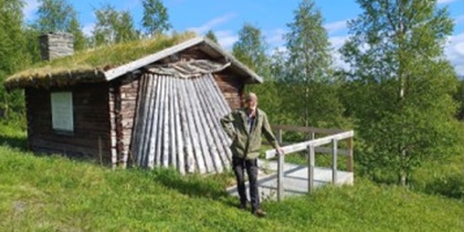 En person står framför en liten timmerstuga med grästak i en grönskande skogsmiljö.