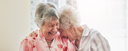 Två äldre kvinnor sitter nära varandra och skrattar tillsammans i ett ljust rum.