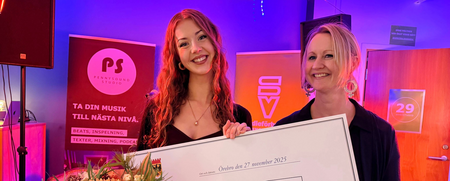 Två personer håller en stor prischeck och en blombukett på en scen med färgglad belysning.