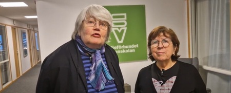 Två äldre kvinnor står inomhus framför en grön skylt med text, båda tittar mot kameran.