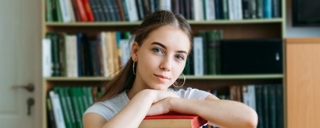 En ung kvinna sitter vid ett bord i ett bibliotek med en hög böcker framför sig.