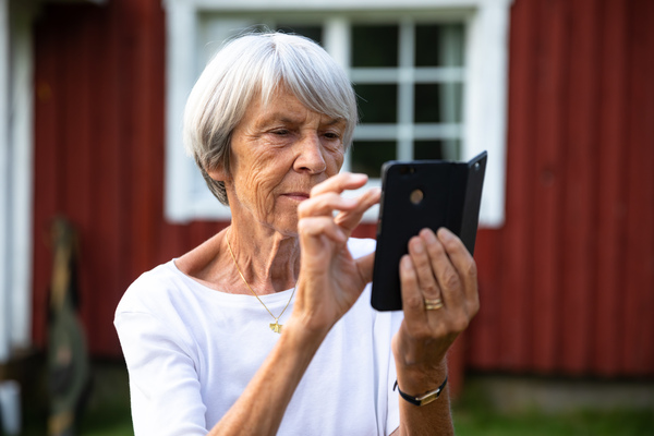Äldre kvinna med grått hår använder en smartphone utomhus framför ett rött trähus.