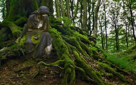 Staty av en sittande person täckt av mossa vid rötter i en grön skog.