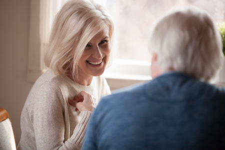 En leende kvinna med ljust hår samtalar med en äldre man vid ett fönster i dagsljus.