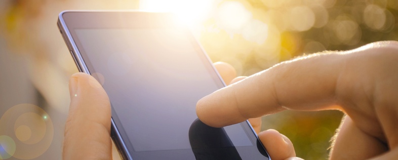 En hand håller en smartphone och en finger rör vid skärmen utomhus i solljus.