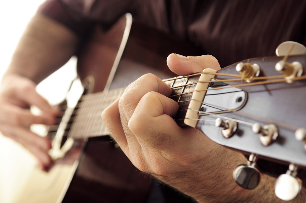 Närbild av en person som spelar akustisk gitarr, fokus på vänster hand på gitarrens greppbräda.