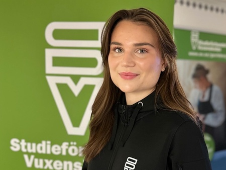 Ung kvinna står framför en grön skylt med logotyp och texten Studieförbundet Vuxenskolan.