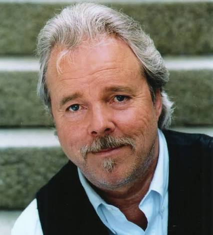 Person med grått hår och svart väst står framför en trappa utomhus.