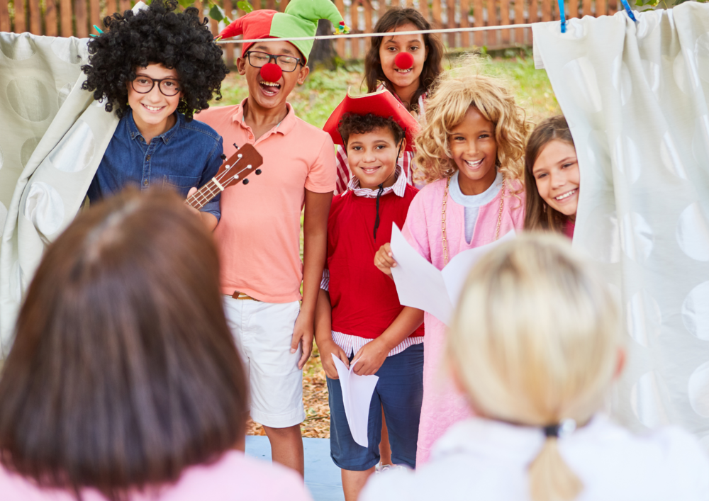 Barn utklädda till clowner och andra figurer uppträder på en utomhusteater inför två åskådare.
