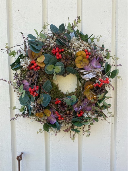 En dörrkrans av torkade blommor, bär, löv och kottar hänger på en vit trävägg.