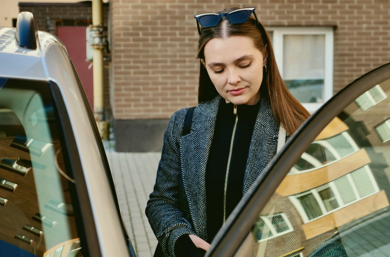 Kvinna med solglasögon öppnar bildörr utomhus vid tegelvägg.