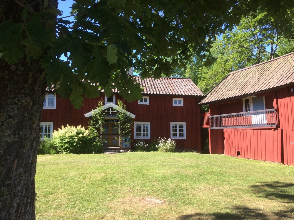 Röd trähusgård med vita fönster, grön gräsmatta och träd i förgrunden en solig dag.