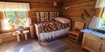 Ett mysigt rum i en timmerstuga med enkel säng, träbord, stol och fönster med blommor och gardiner.