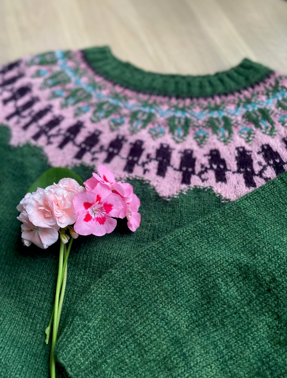 Grön stickad tröja med rosa och lila mönster, dekorerad med rosa blommor ovanpå.