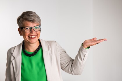 Leende kvinna med glasögon och kort grått hår, bär grön topp och ljus kavaj, håller ena handen uppåt.