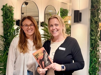 Två kvinnor står och ler i ett modernt kontorsrum, en håller en bok med titeln 'Succé Syster'.
