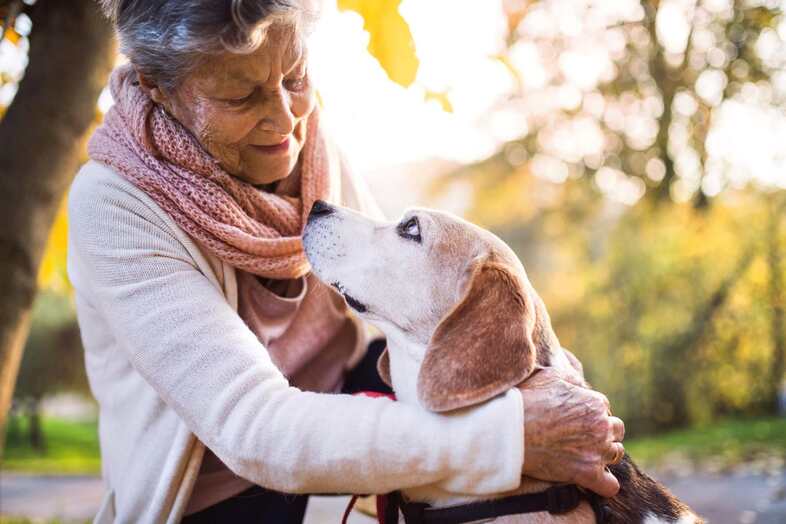 En äldre kvinna sitter på huk och håller en arm om en hund. Kvinnan och hunden tittar varandra i ögonen.