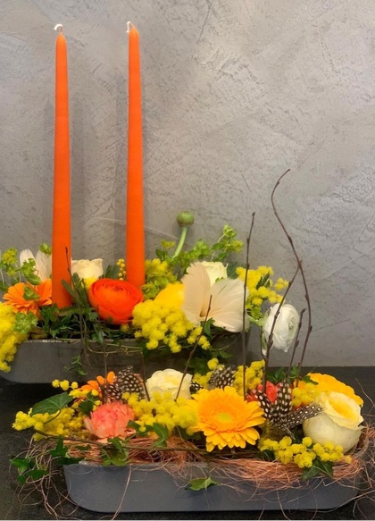 Två blomsterarrangemang med orange ljus, gula och vita blommor samt dekorativa fjädrar på ett bord.