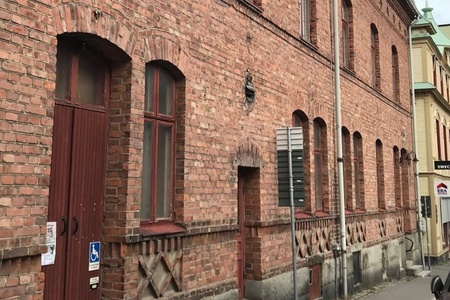 Röd tegelbyggnad med höga fönster och dörrar längs en smal stadsgata, parkerad bil till höger.