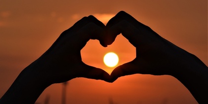 Två händer formar ett hjärta framför en solnedgång med solen i mitten av hjärtat.