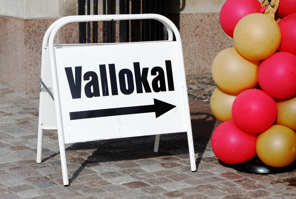 Skylt med texten 'Vallokal' och en pil åt höger, bredvid röda och gula ballonger på stenlagd mark.