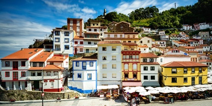 Färgglada hus i flera våningar står tätt intill varandra vid en kullerstensgata i en kuststad med grönskande backe bakom.