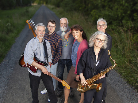 Sex vuxna personer står på en grusväg utomhus, två håller musikinstrument, alla ler mot kameran.