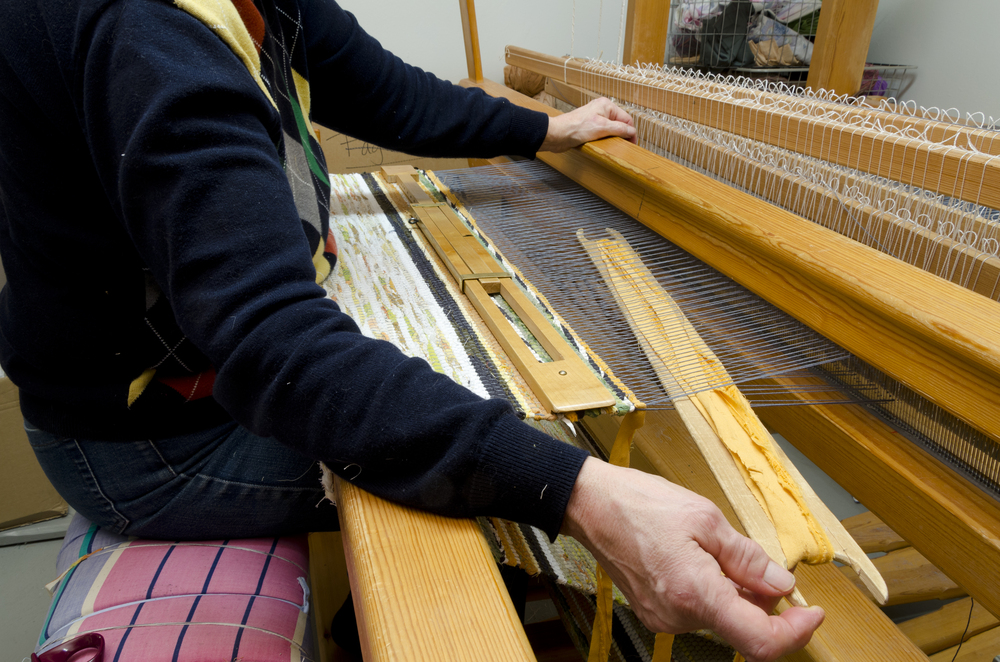 En person väver tyg på en stor vävstol i ett ljust rum. Händerna arbetar med trådarna.