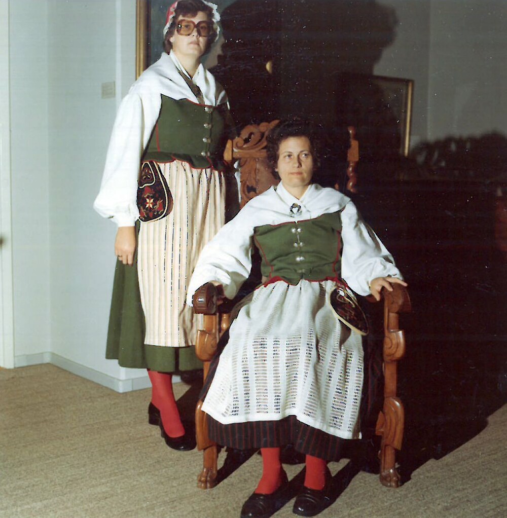 Två personer i traditionella folkdräkter, en sittande och en stående, inomhus vid en trästol.