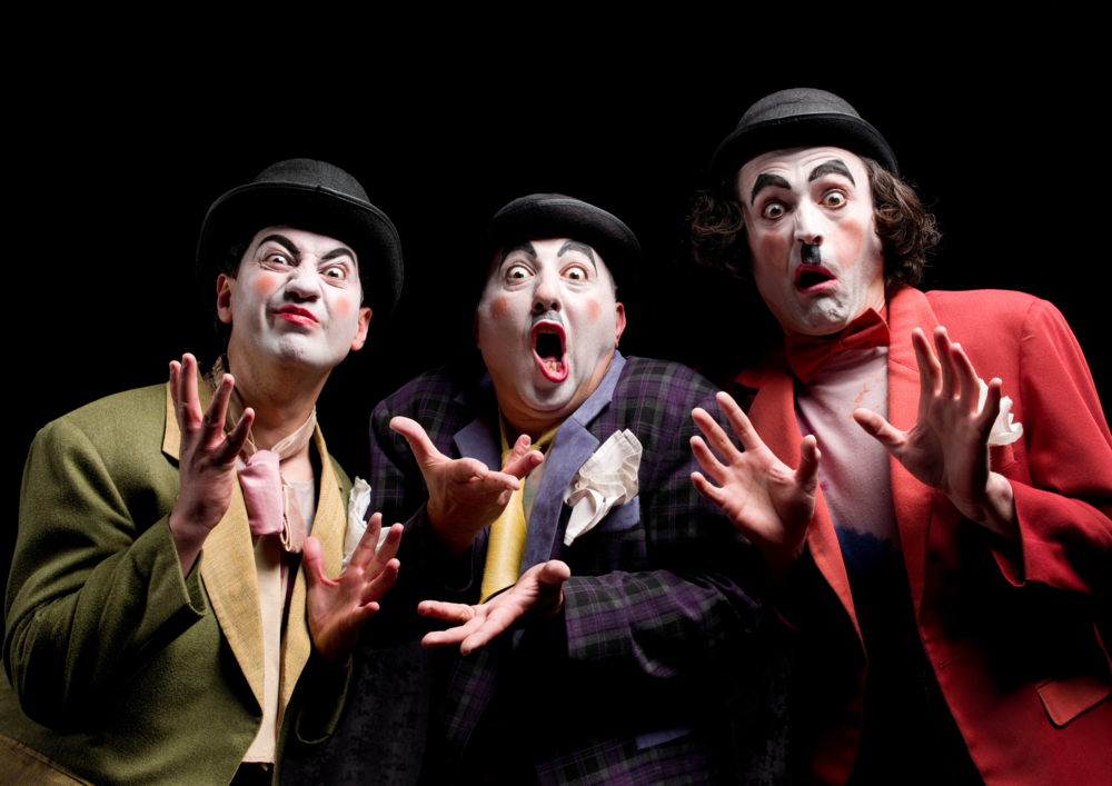 Tre clowner med vit ansiktsmålning och färgglada kläder ser förvånade ut mot svart bakgrund.