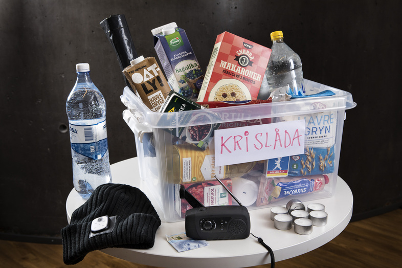 En plastlåda märkt 'KRISLÅDA' fylld med mat, vattenflaskor och nödvändiga saker står på ett runt bord.