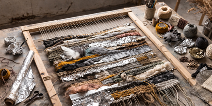 Vävram med pågående vävprojekt i naturfärger och folie, omgiven av garn, saxar och hantverksmaterial på ett bord.