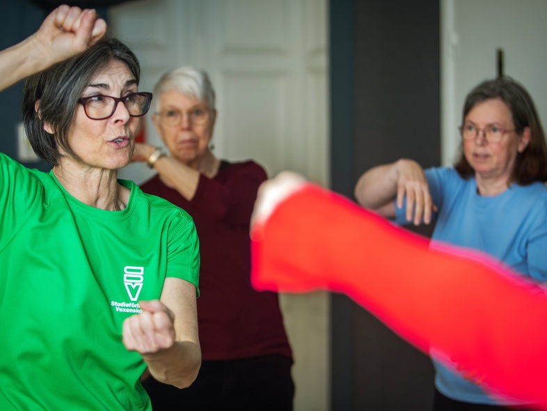 Tre äldre kvinnor deltar i en tränings- eller självförsvarsklass inomhus, en i grön tröja leder övningen.