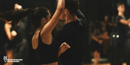 Två personer dansar tillsammans i en dansstudio med andra par i bakgrunden.