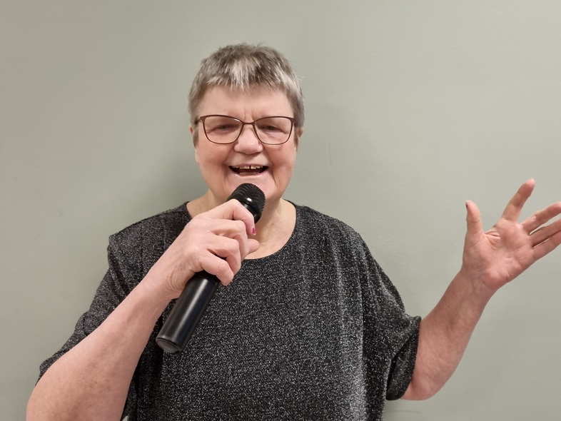 Person med grått hår håller en mikrofon och gestikulerar mot en ljusgrå vägg.