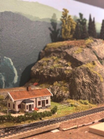 Modelljärnväg med ett litet hus, järnvägsspår och berg i bakgrunden.