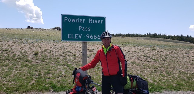 Cyklist i röd jacka står vid en skylt för Powder River Pass, höjd 9666 fot, med cykel och packning.