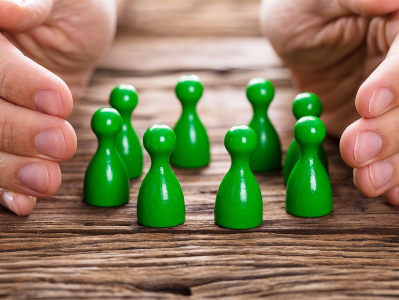 Två händer skyddar en grupp gröna spelpjäser på ett träbord.