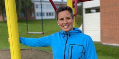 Person i blå jacka står vid klätterställning på en lekplats utomhus, ler mot kameran.