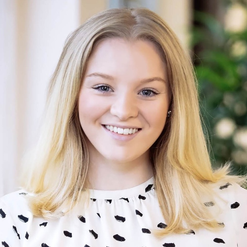 Ung kvinna med blont hår och vit blus med svarta prickar ler mot kameran inomhus.