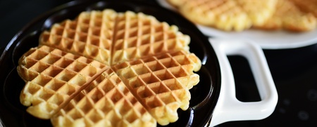 Nybakade hjärtformade våfflor i ett våffeljärn med fler våfflor på en tallrik i bakgrunden.