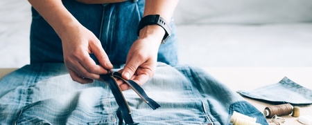 Person arbetar med att sy om ett par jeans, med dragkedja, måttband och sytråd på bordet.